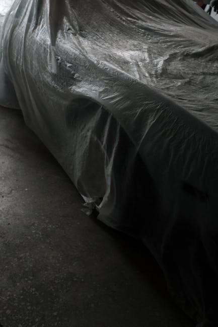 A classic vehicle hidden under a plastic sheet in a dimly lit garage, emphasizing its preservation.